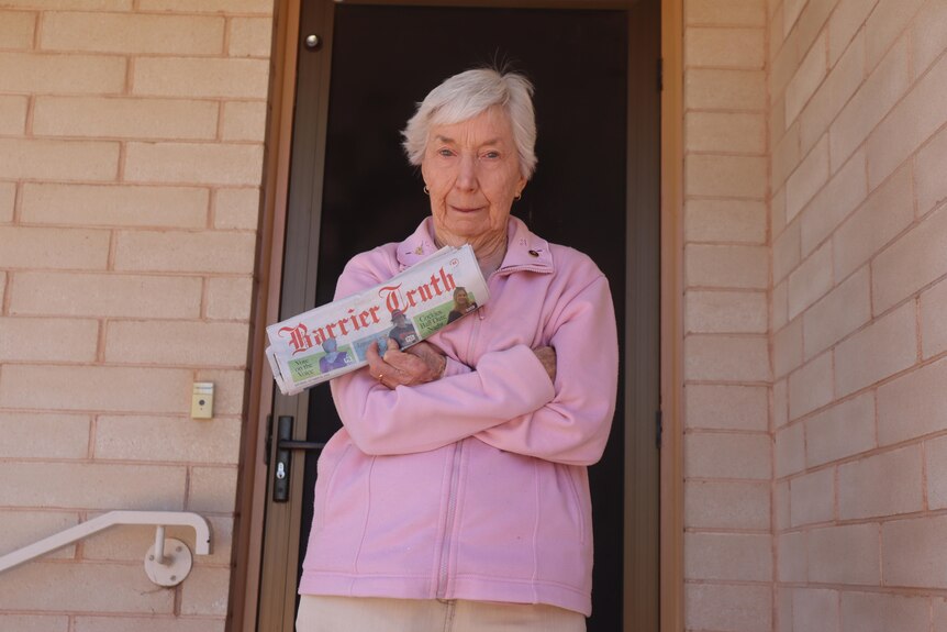 An older woman wearing a pink cardigan holding a newspaper with her arms crossed in front of a door.