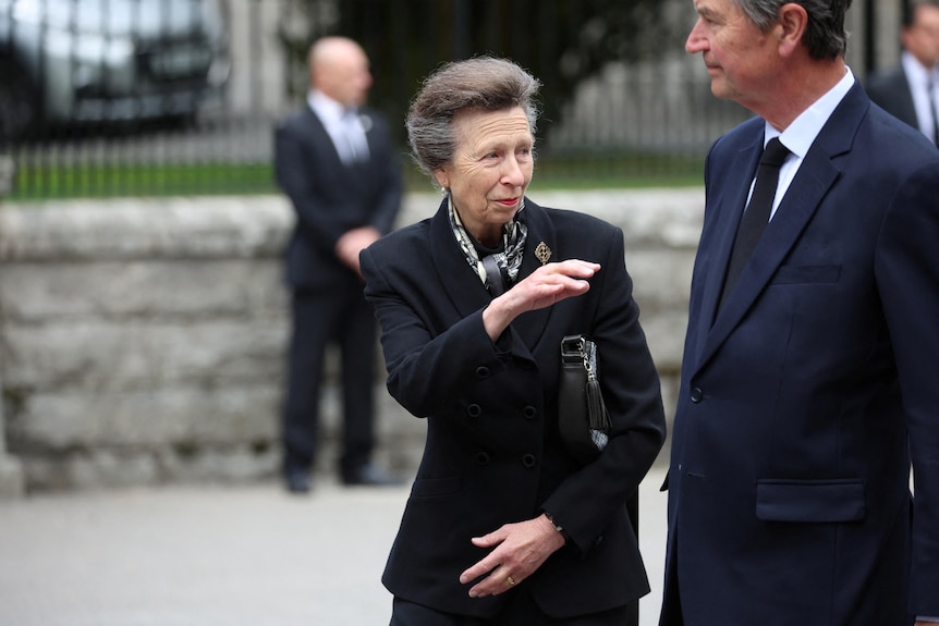 Princess Anne waves a hand as a man in suit stands next to her. 