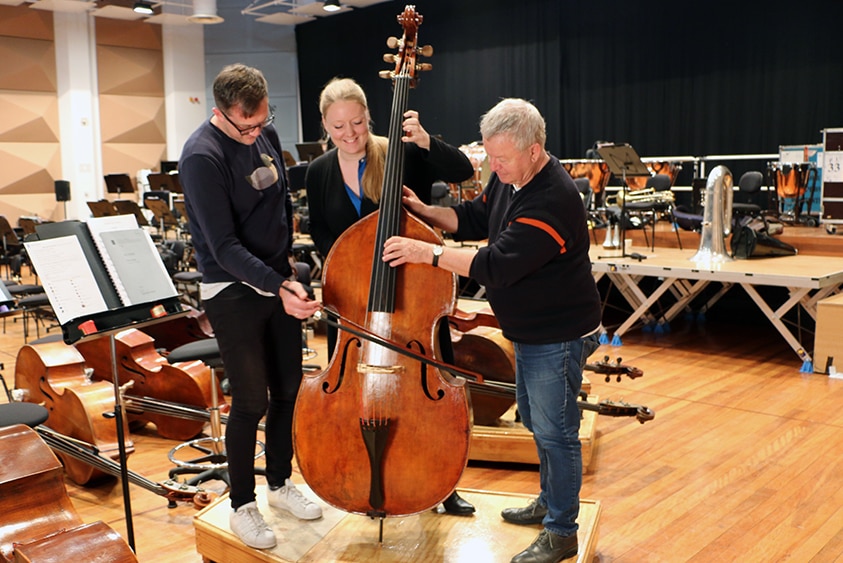 Three double bass players stand around a single instrument, pretending to play it together.