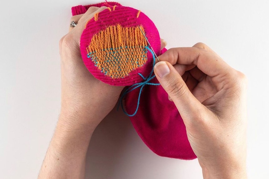 A woman's hand holds a bright pink sock and her other hand darns it with a needle and orange thread