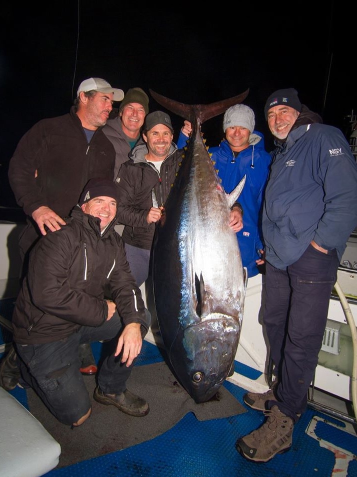 Six men on a boat smiling and holding a very large tuna
