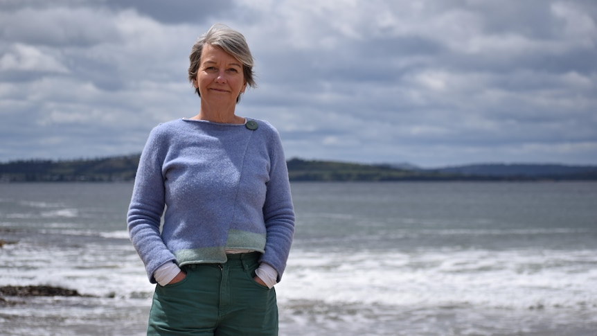 A woman stands on a beach, half-smiling, she has grey hair, wears blue woolen top and dark green pants