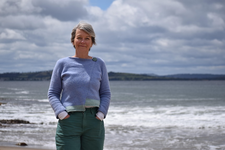A woman stands on a beach, half-smiling, she has grey hair, wears blue woolen top and dark green pants