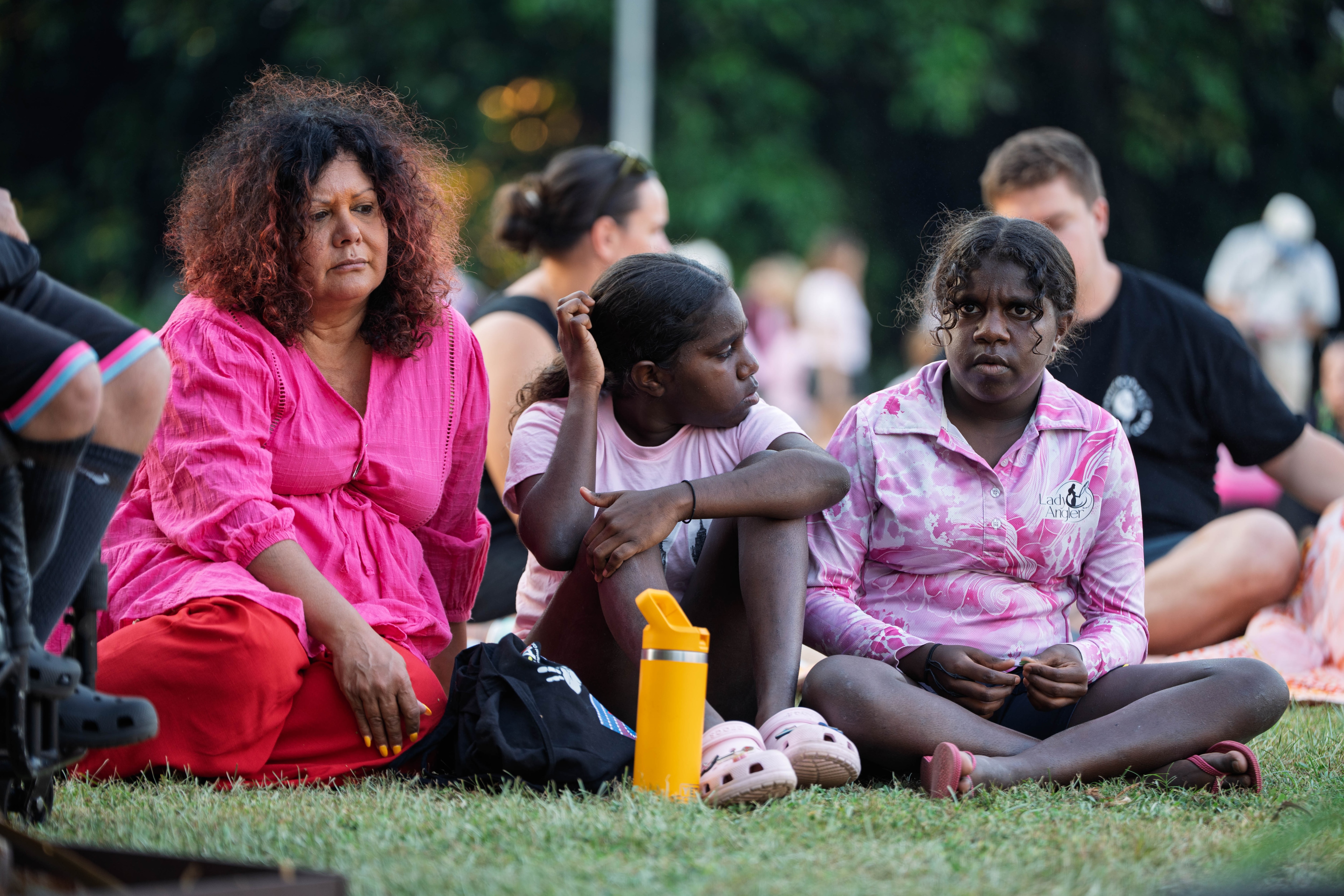An Indigenous woman in a pink dress sitting on grass alongside two teens, they are all wearing serious facial expressions.