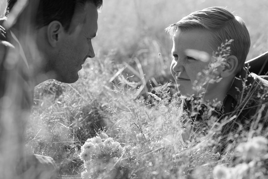 A black and white image of a boy in 60s-style clothing lying in the grass, across from a middle-aged man