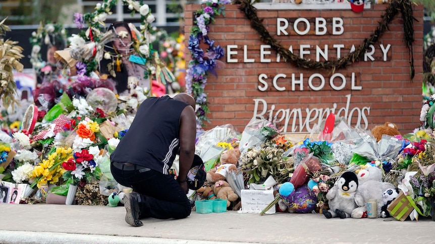  A black man kneels in front of a brick wall with ROBB ELEMENTARY SCHOOL. Flowers, toys and cards cover it. 