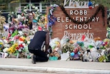  A black man kneels in front of a brick wall with ROBB ELEMENTARY SCHOOL. Flowers, toys and cards cover it. 