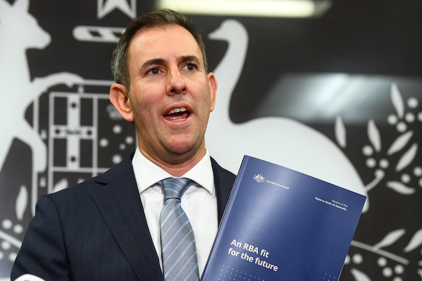 A man wearing a suit stands in front of the Australian emblem holding  a report.