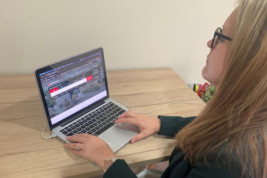 A photo from behind , showing the side of Megan sitting at a table with her laptop, staring at the screen. 