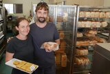 Husband and wife stand at the front of their bakery, holding apple pies and a chelsea bunn