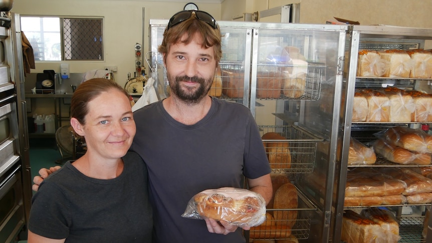 A man and a woman stand at the front of their bakery, holding apple pies and a chelsea bunn