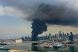 You view the charred cityscape of Beirut from its port as black smoke rises from the entrance to the port.