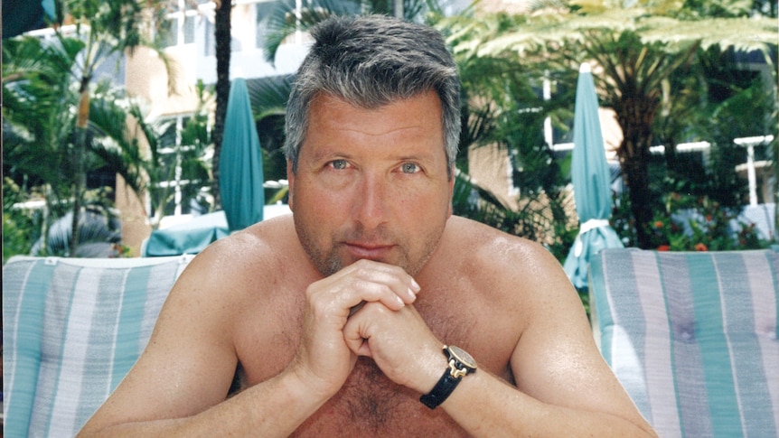 A man sits resting his chin on his hands with deckchairs, umbrellas and fern trees in the background.