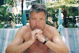 A man sits resting his chin on his hands with deckchairs, umbrellas and fern trees in the background.