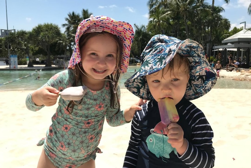 A four year old girl eats a chocolate ice-cream on beach next to 3 year old boy with rainbow one.