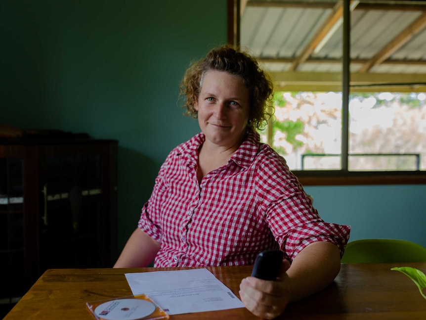 A woman in a checked shirt, sits at a table holding a phone, with a piece of paper and CD in front of her. Window in background.