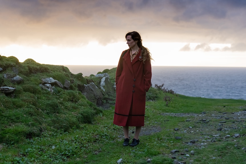 A troubled-looking woman in her late 30s in a 20s-style red coat stands in the Irish countryside on an overcast day.