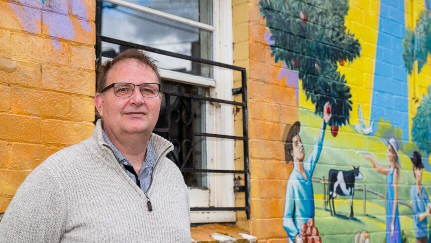 Peter, 55, stands wearing a grey jumper in front of a colourful wall.