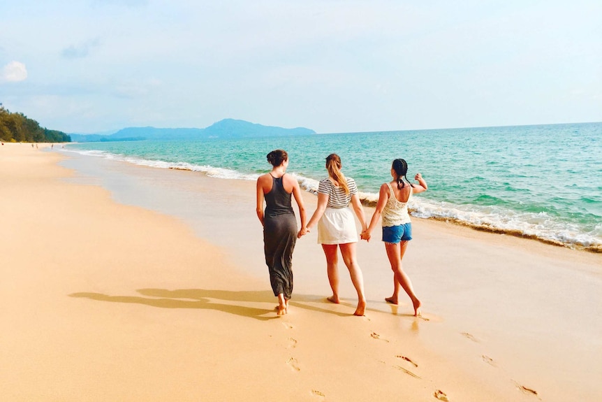 Three women walk along a beach holding hands.