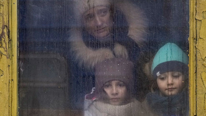 Two young children wearing beanies and coats look through a dirty train window with a worried woman in the background.