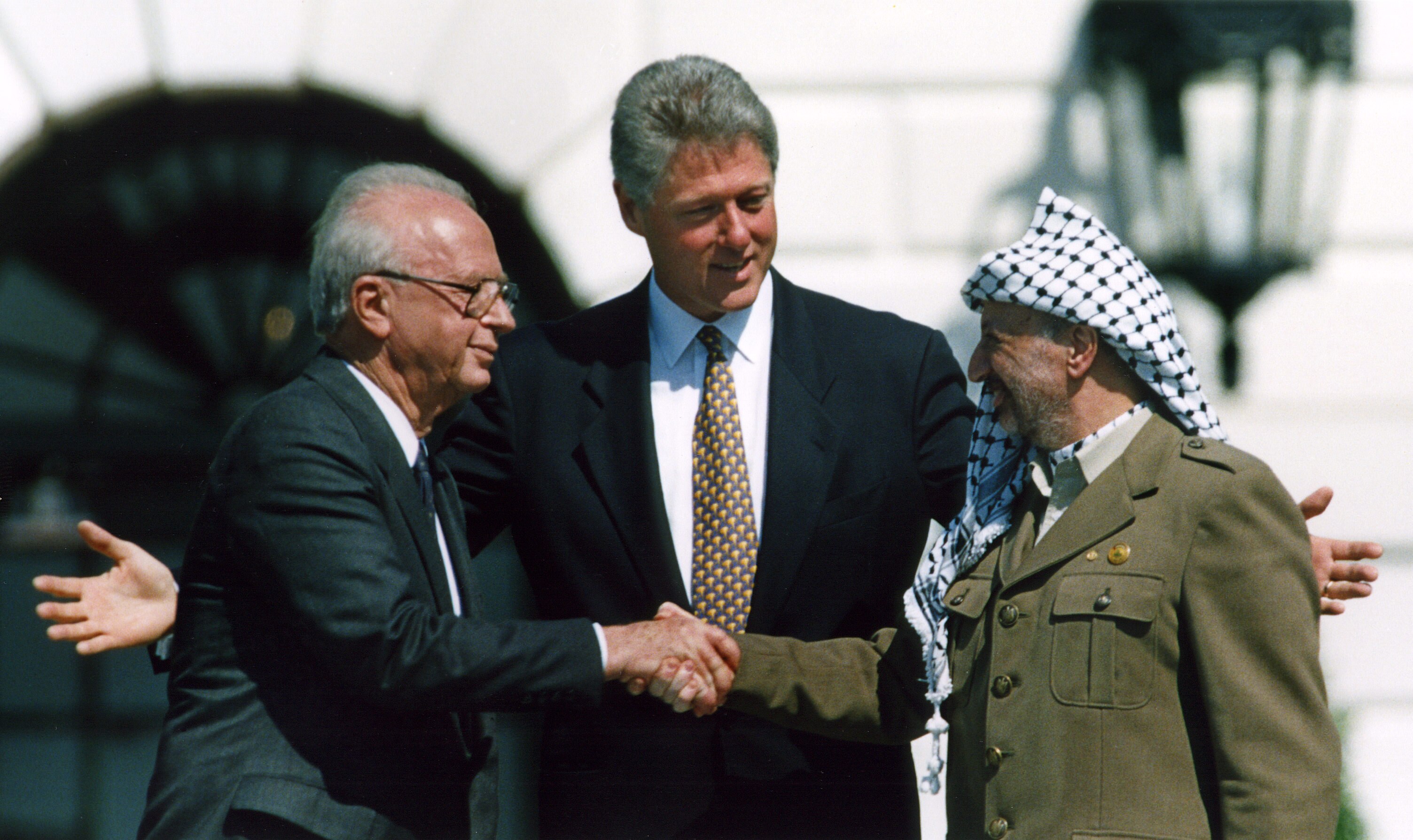 Two men shake hands as one man stands in between them.
