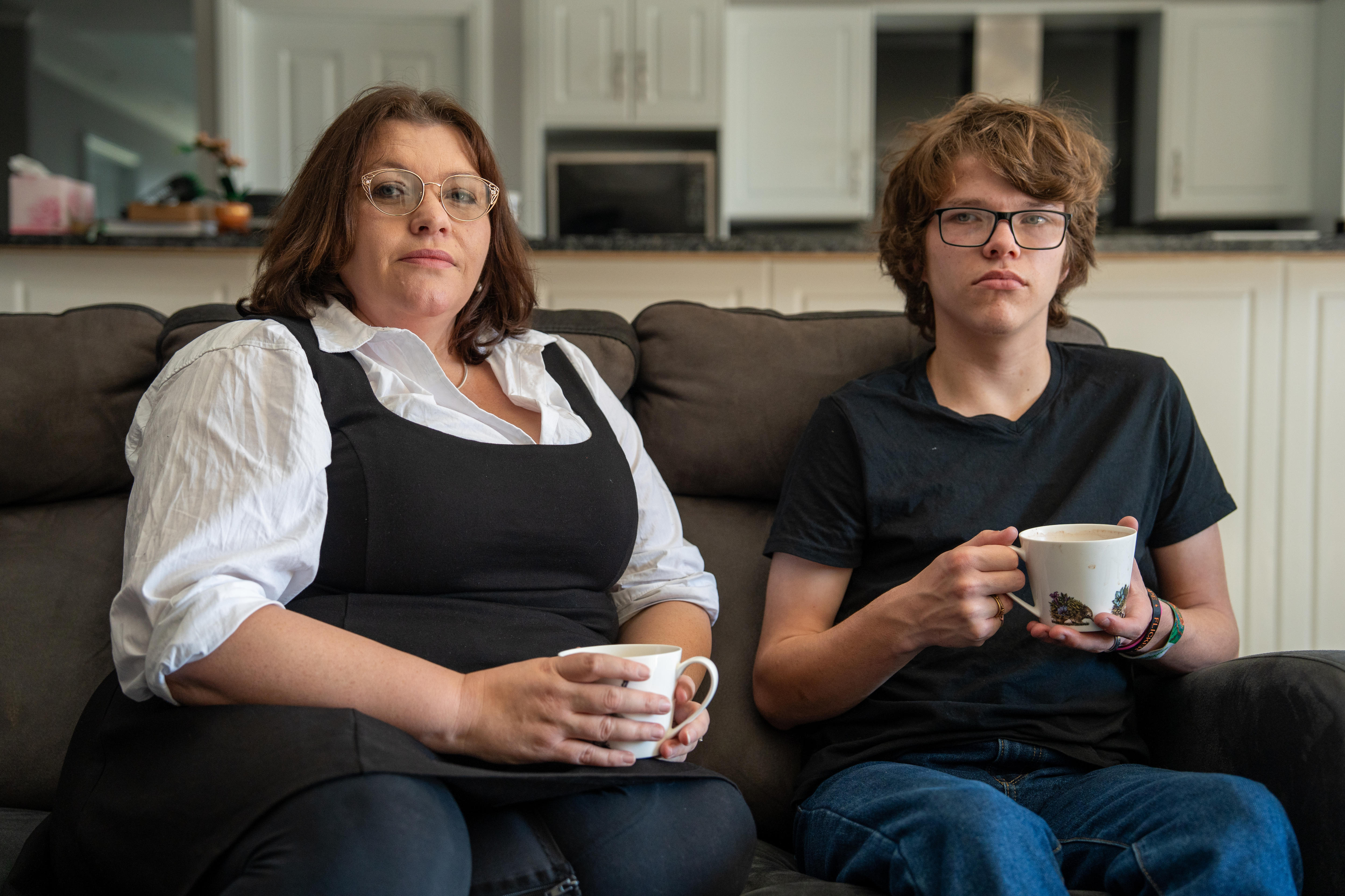 A woman and a teenage boy each holding a mug while sitting on a couch with a kitchen in the background