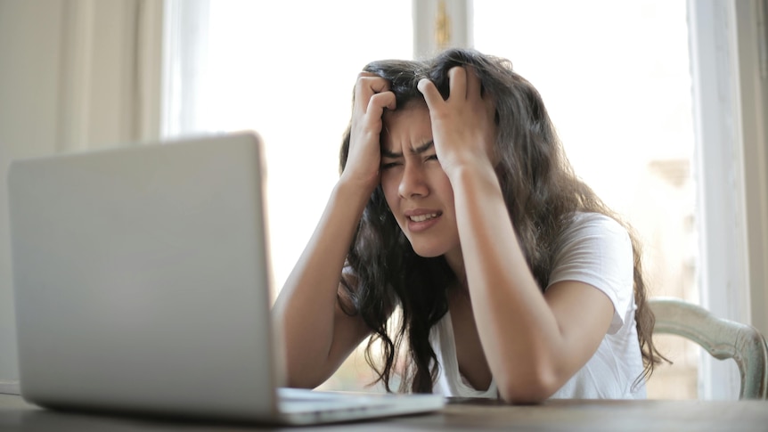 Young girl with long hair wearing a white t-shirt looking at a laptop holding her head looking stressed  