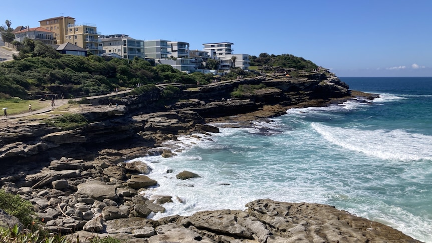 A cliff with houses on top overlooks a bay of rocks with waves splashing up the side. 