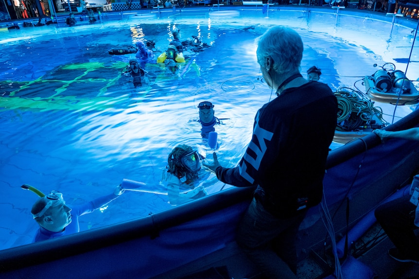 James Cameron stands at the edge of a large pool in which actors swim in scuba masks