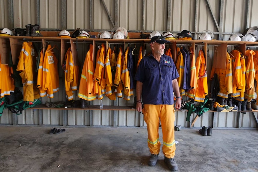 A member of the Woodgate Rural Fire Brigade stands in a shed, with fire gear hanging up behind him on the wall.