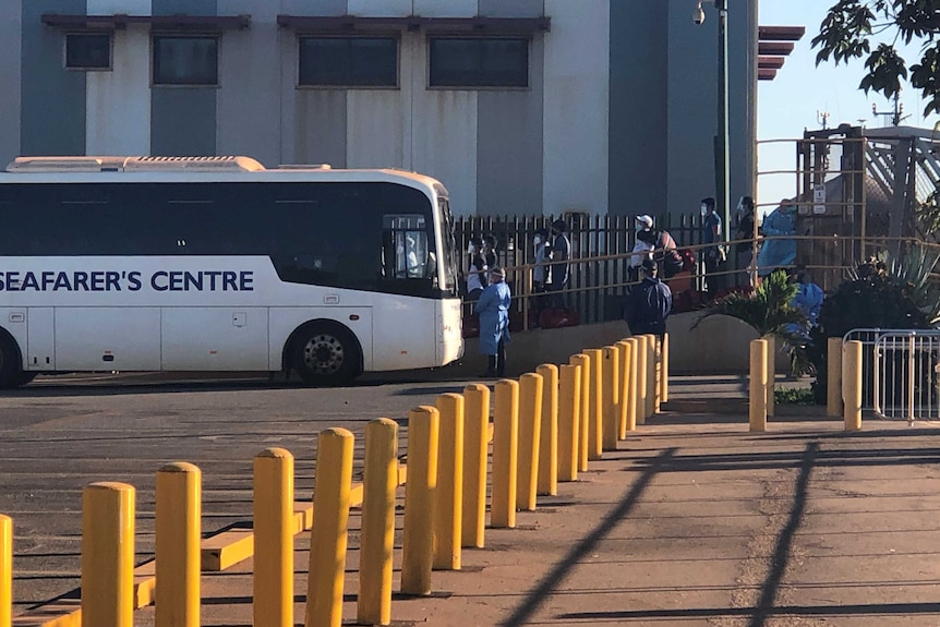 A bus at the end of a gangway at a shipping port, with numerous people in masks walking.