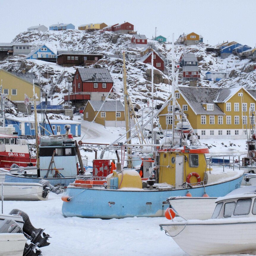 Fishing boats sit in the harbour of Uummannaq, with houses on a snow-covered slope behind.