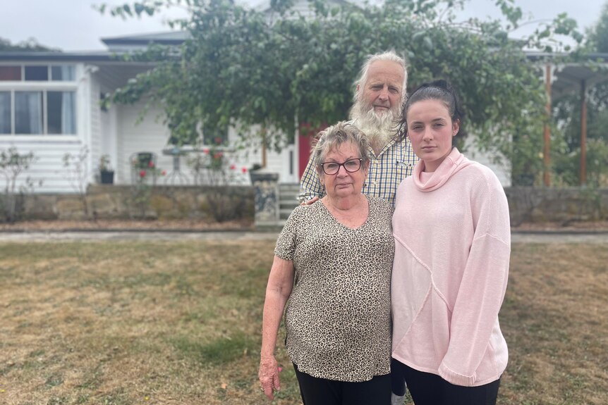 Two grandparents with their 21 year old granddaughter, standing outside their home. 