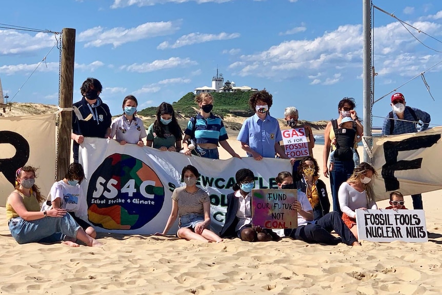 A group of 17 people hold signs as they stand and sit on the beach in Newcastle, urging for action against climate change.