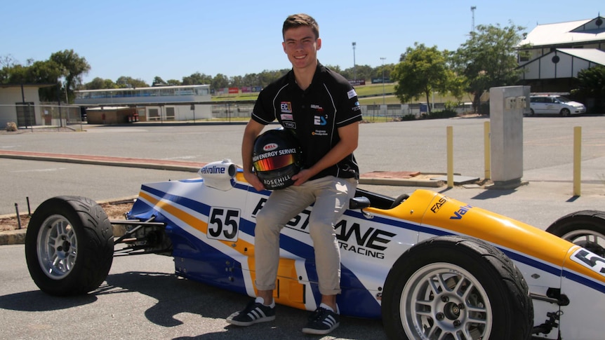 Calan sits on top of a yellow, blue and white racing car holding a racing helmet.