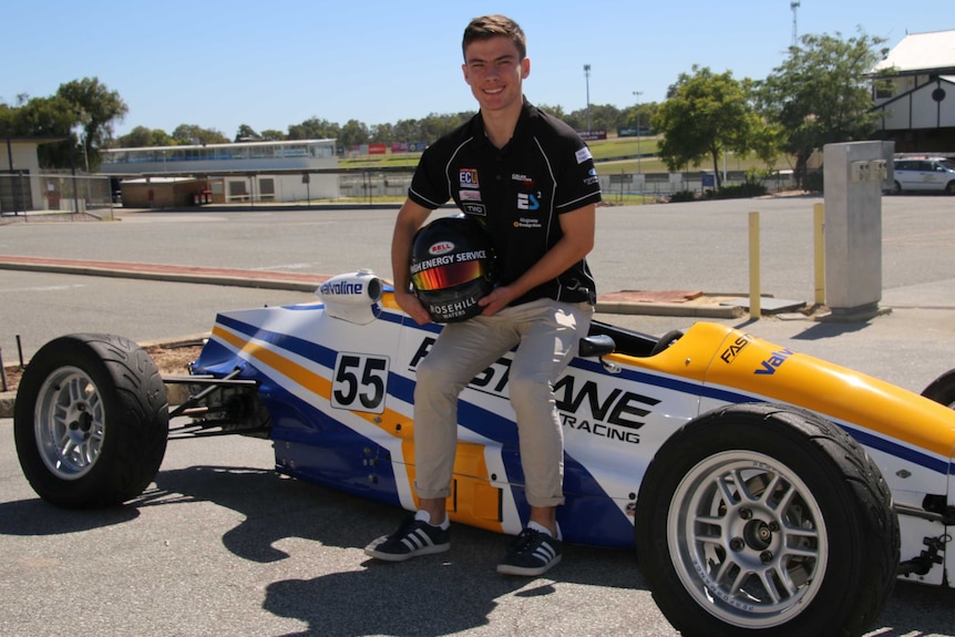 Calan sits on top of a yellow, blue and white racing car holding a racing helmet.