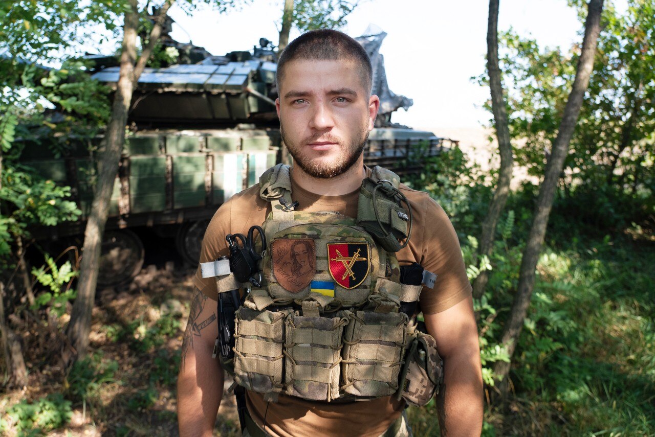 A man wearing combat clothing looks at the camera with a serious expression.