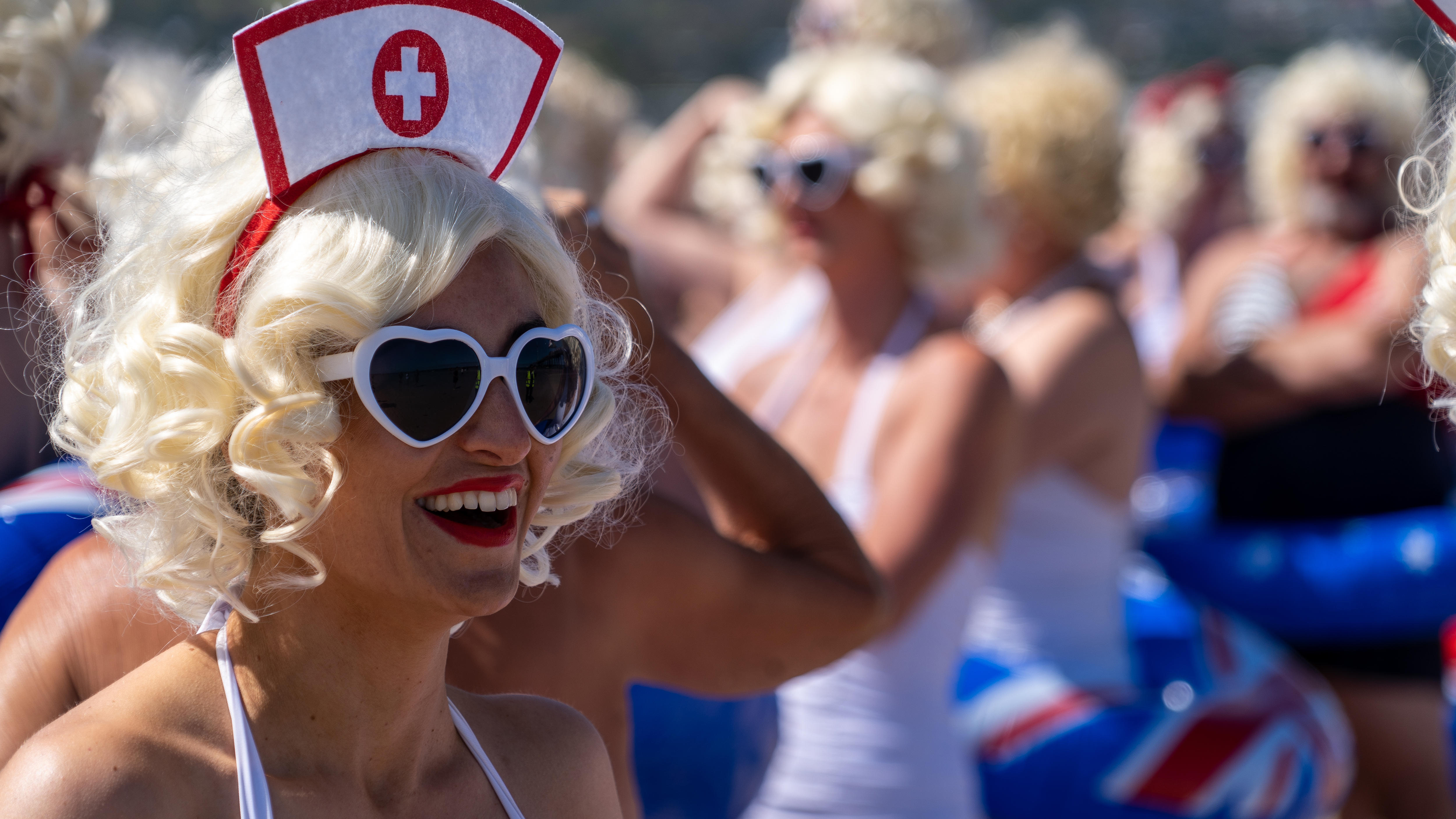 A woman wearing a blonde curly wig, white heart-shaped sunglasses and a nurses hat smiles while looking off camera