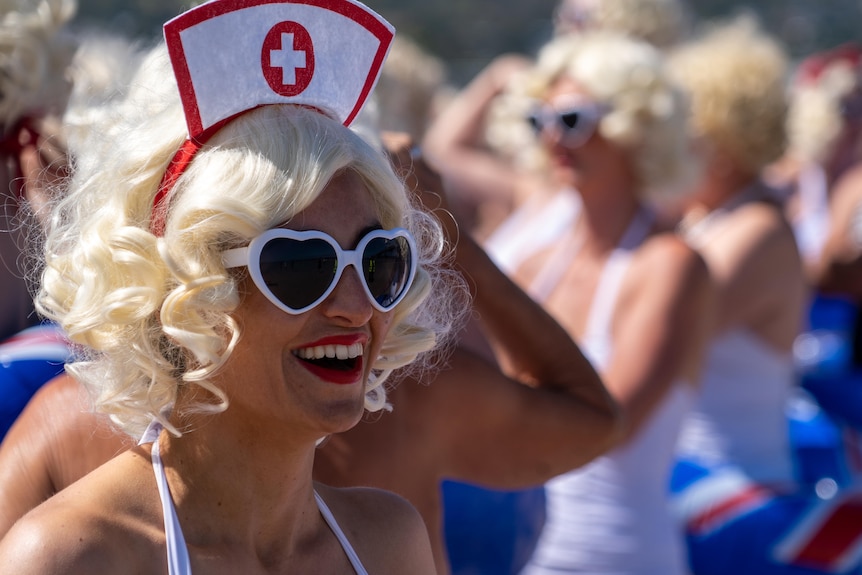 A woman wearing a blonde curly wig, white heart-shaped sunglasses and a nurses hat smiles while looking off camera