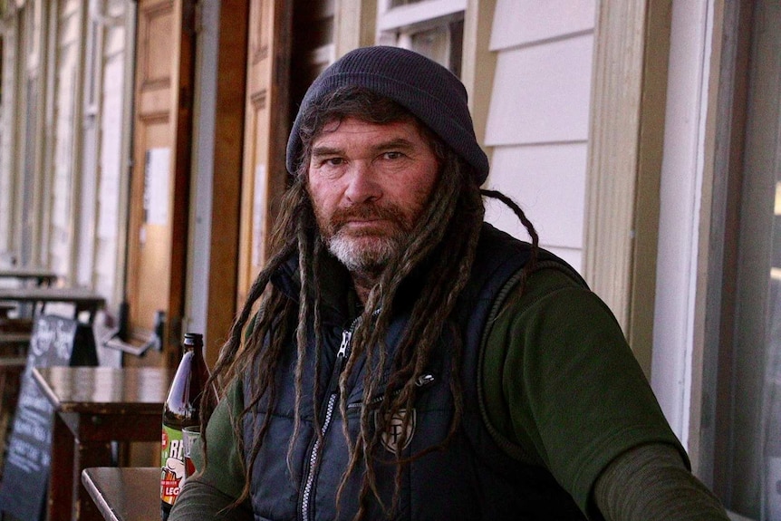 A man with dreadlocks and a beanie sits out the front of the pub with a beer.