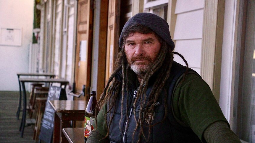 A man with dreadlocks and a beanie sits out the front of the pub with a beer.