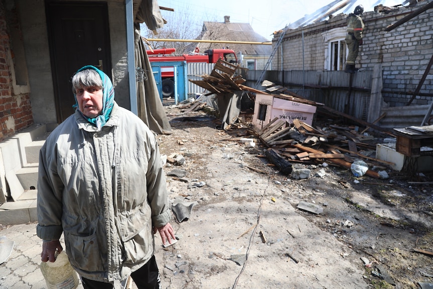 Kobieta w średnim wieku ubrana w zieloną chustę na głowie stoi przed zniszczeniami wojennymi w osiedlu mieszkaniowym