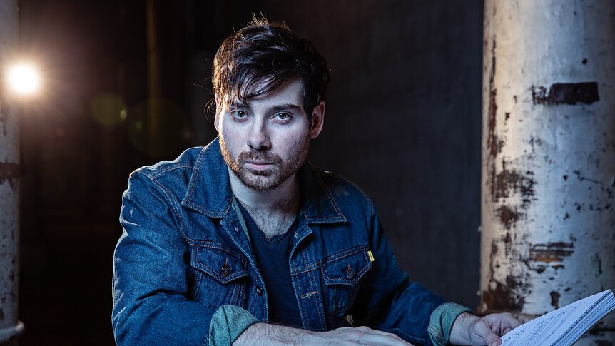 Man wearing denim jacket looking to camera and sitting in darkly lit industrial space with libretto in front of him.