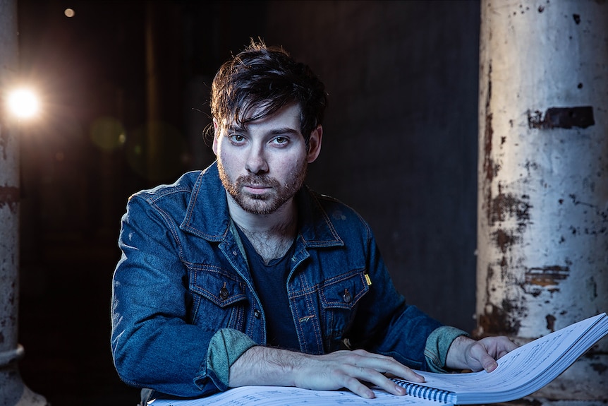 Man wearing denim jacket looking to camera and sitting in darkly lit industrial space with libretto in front of him.