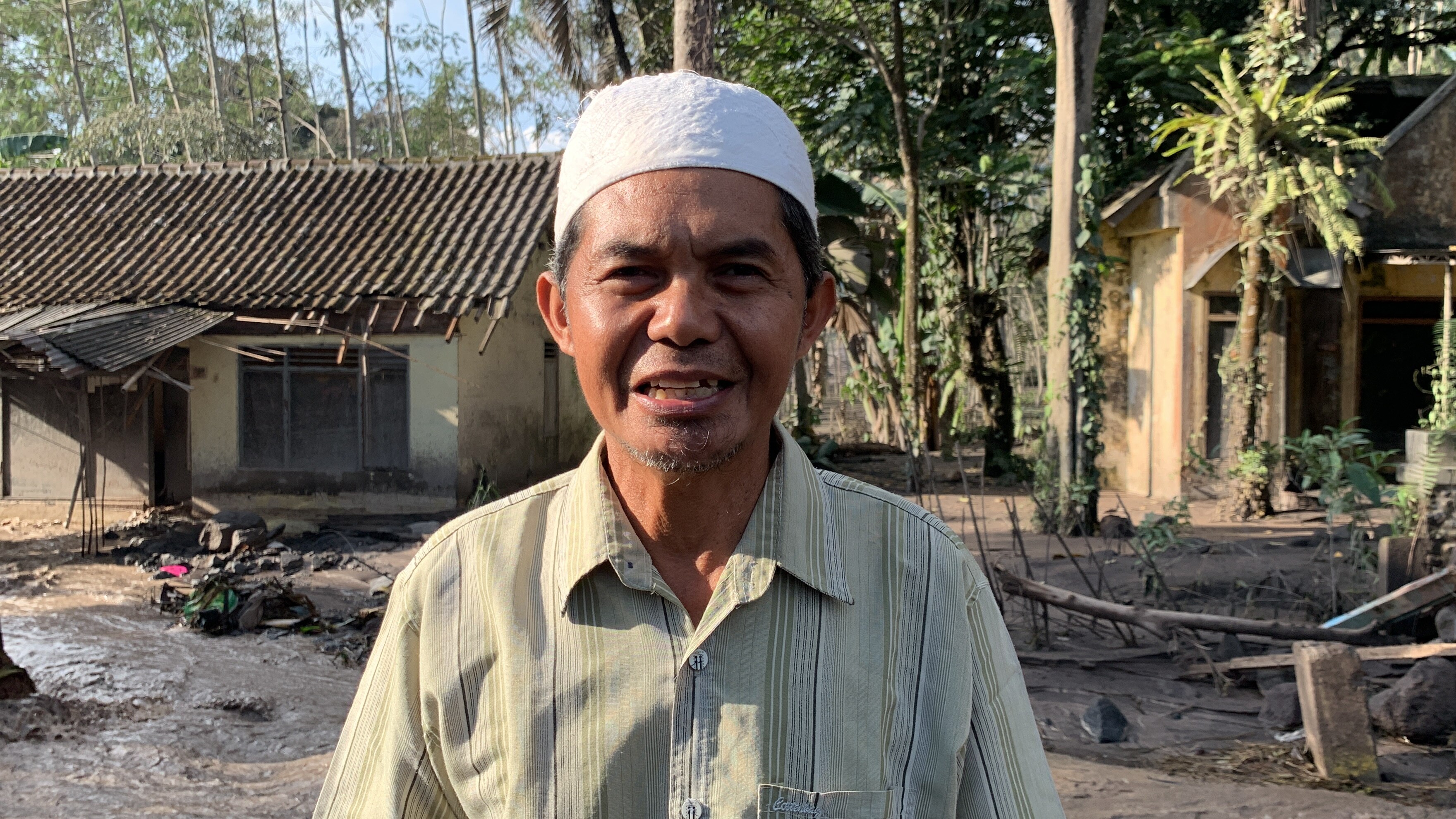 A man with a white skullcap in front of his damaged house.
