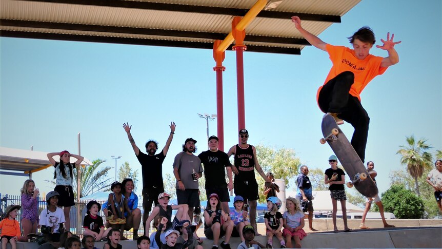 A man in an orange shirt is mid-air on a skateboard while a crowd of mostly children watch on behind. 