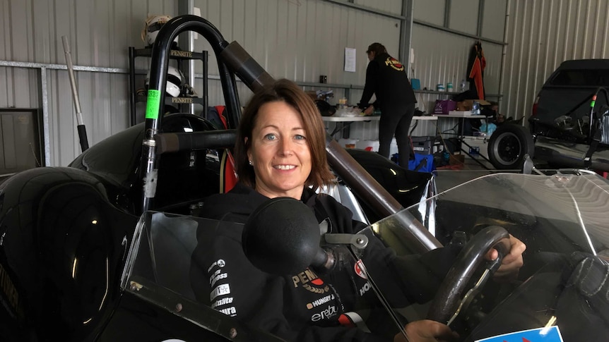 Female drive sits in black race car with hands on the steering wheel.
