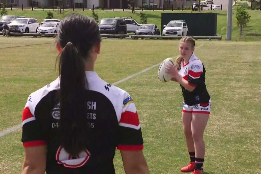 A female rugby league player holds the ball in her hands, preparing to pass to teammate in training.