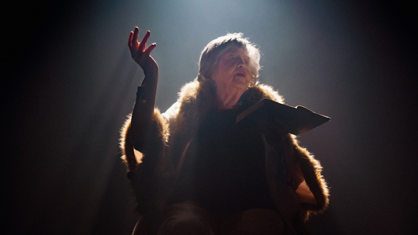 An older woman sitting a under a spot stage light holding a book and performing.