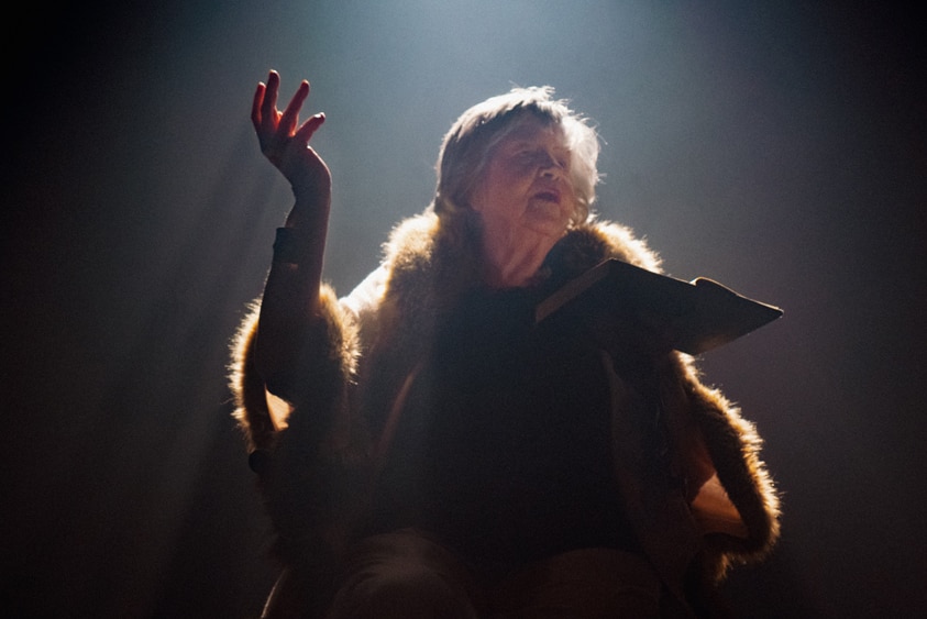 An older woman sitting a under a spot stage light holding a book and performing.